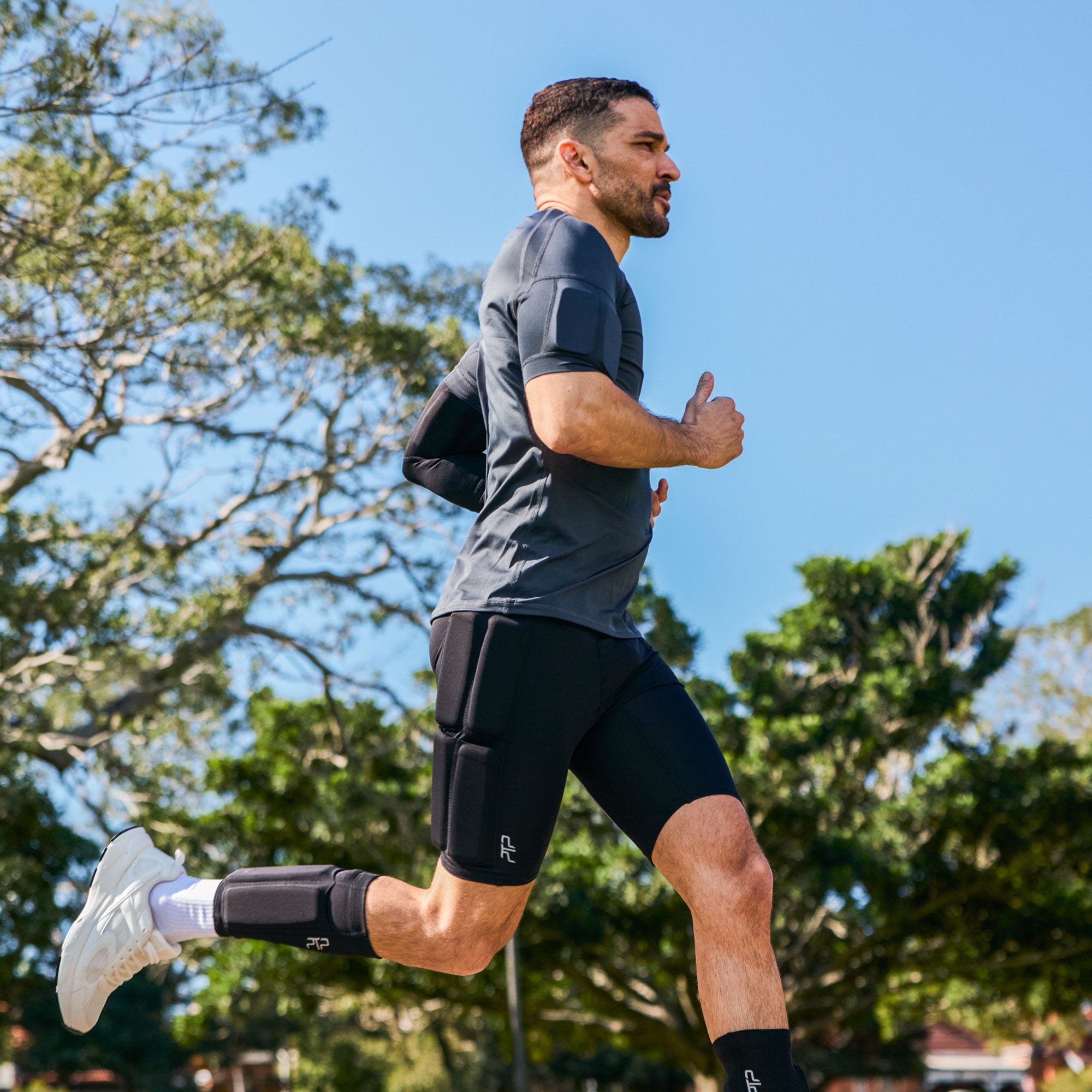 A man jogging outdoors wearing integrated strengthwear. Dark gray athletic t-shirt, black running shorts with a built-in knee pad, a black knee sleeve, and white running shoes, with trees and a blue sky in the background.