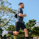 A man jogging outdoors wearing integrated strengthwear. Dark gray athletic t-shirt, black running shorts with a built-in knee pad, a black knee sleeve, and white running shoes, with trees and a blue sky in the background.