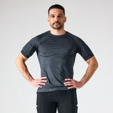 A man wearing a dark gray short-sleeve athletic PTP compression shirt and black pants, standing with hands on hips against a light gray studio background.